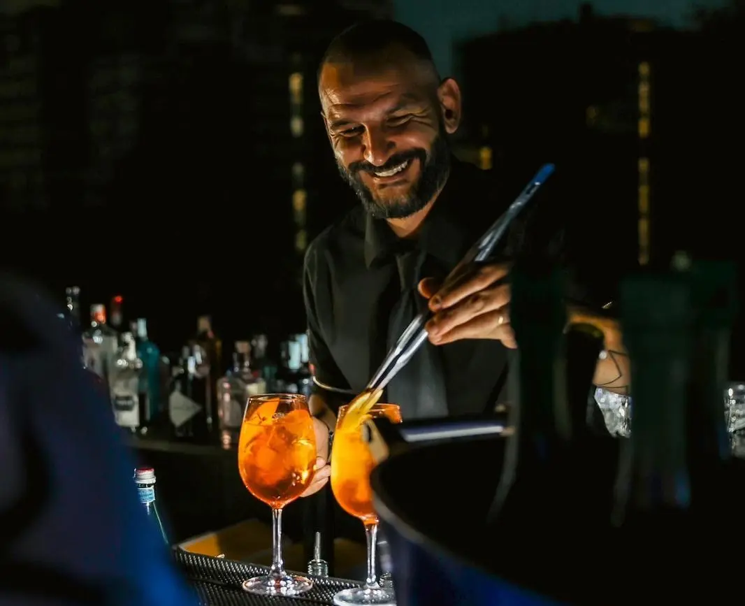Filippo sorride mentre prepara due spritz arancioni al banco bar, in un locale buio con le bottiglie sullo sfondo.