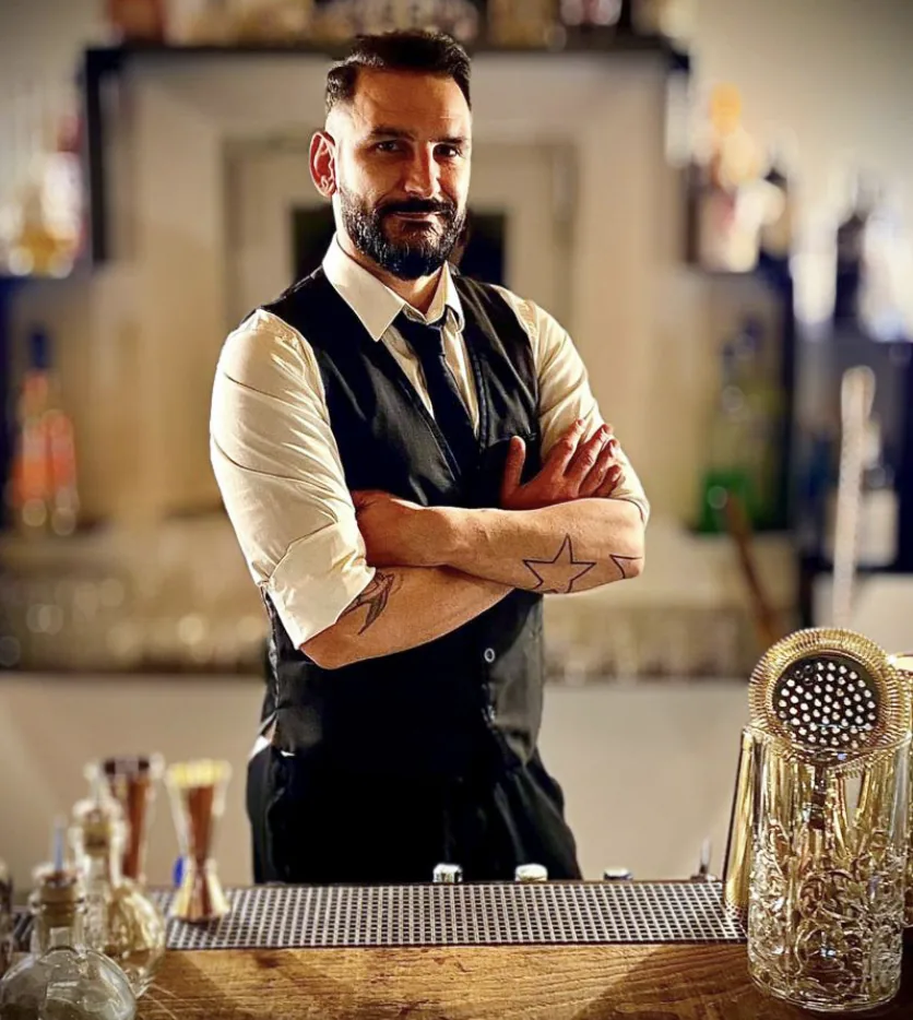 Filippo, con gilet nero, cravatta e camicia bianca con le maniche arrotolate, sta a braccia conserte dietro il banco; in primo piano il tappetino da bar, alcuni contagocce e un miscelatore in cristallo, alle spalle le mensole con le bottiglie.