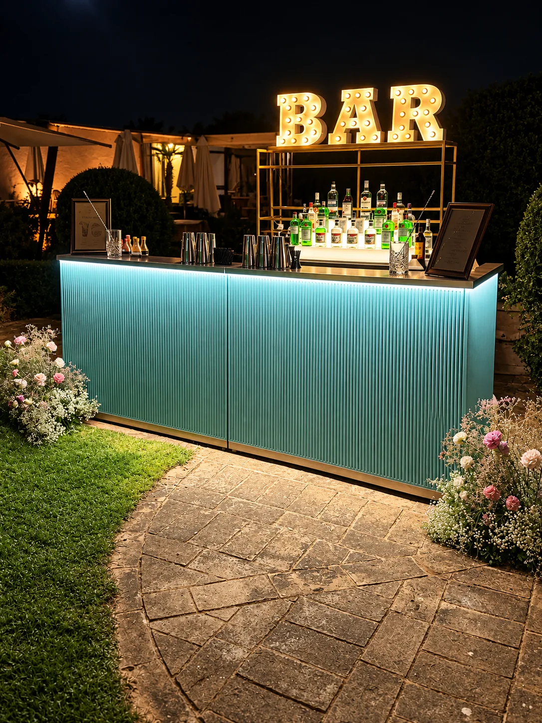 Bancone mobile color tiffany con frontale scanalato, illuminato di sera su un patio di pietra accanto al prato: sopra l'insegna luminosa a lampadine con la scritta BAR, dietro una scaffalatura dorata con le bottiglie, sul piano shaker, bicchieri e due listini cocktail incorniciati, con mazzi di fiori ai lati.