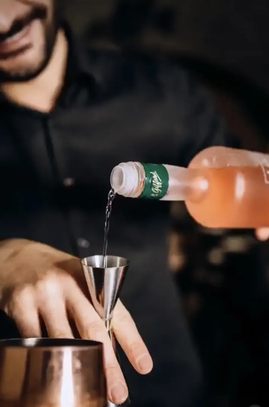 Un bartender versa un cocktail da una bottiglia in un misurino d'acciaio, con uno shaker di rame in primo piano su uno sfondo scuro.