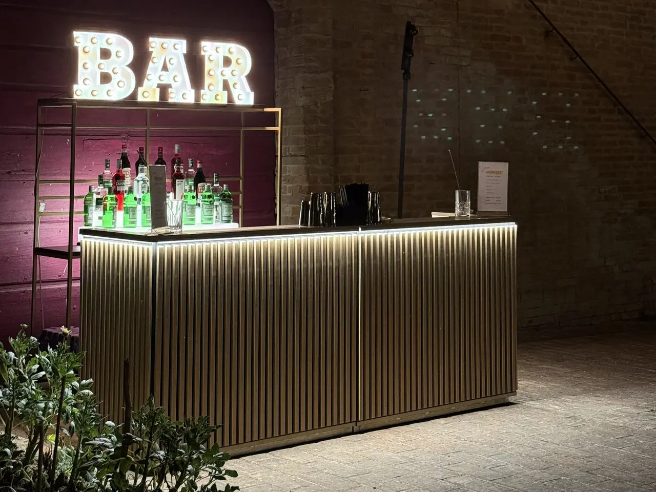 Un bancone mobile con frontale in listelli di legno chiari, illuminato da una striscia LED, con una mensola di bottiglie e l'insegna luminosa BAR, in una corte in pietra di sera.