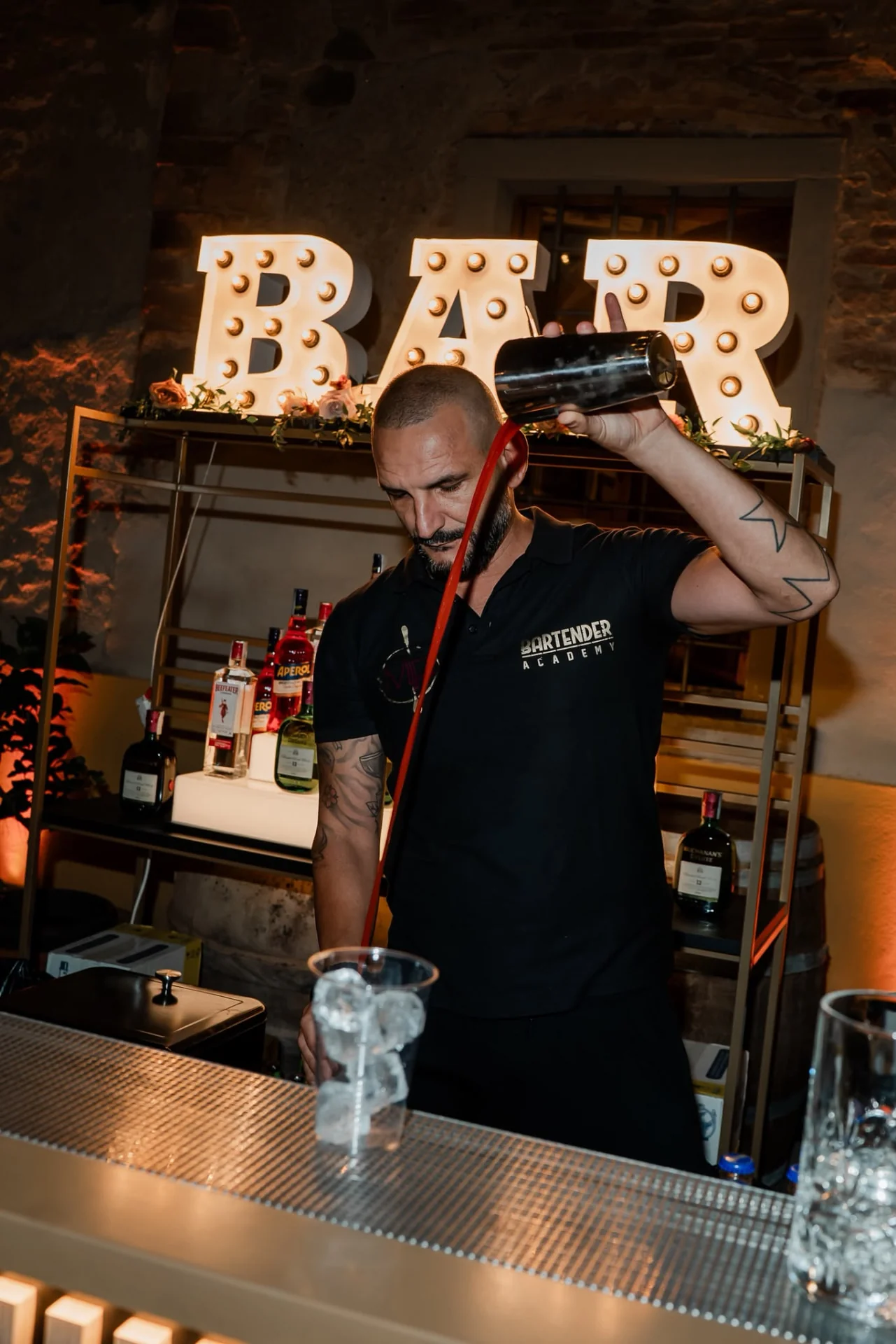 Un bartender dell'Academy versa un cocktail rosso dallo shaker in un bicchiere con ghiaccio, davanti all'insegna luminosa BAR.