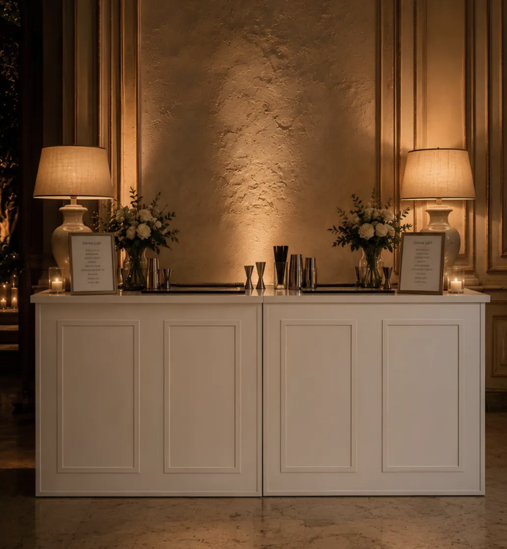 Un bancone bianco con pannelli a cornice, due lampade con paralume acceso, due vasi di rose bianche e shaker al centro, in una sala elegante con pareti in stucco e pavimento in marmo.
