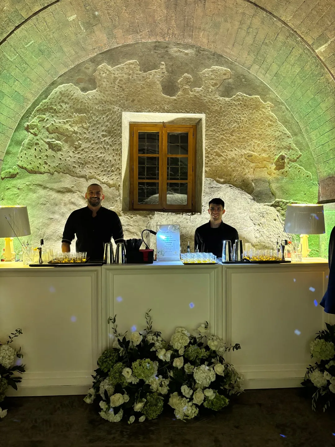 Due bartender sorridono dietro un bancone bianco decorato con fiori.